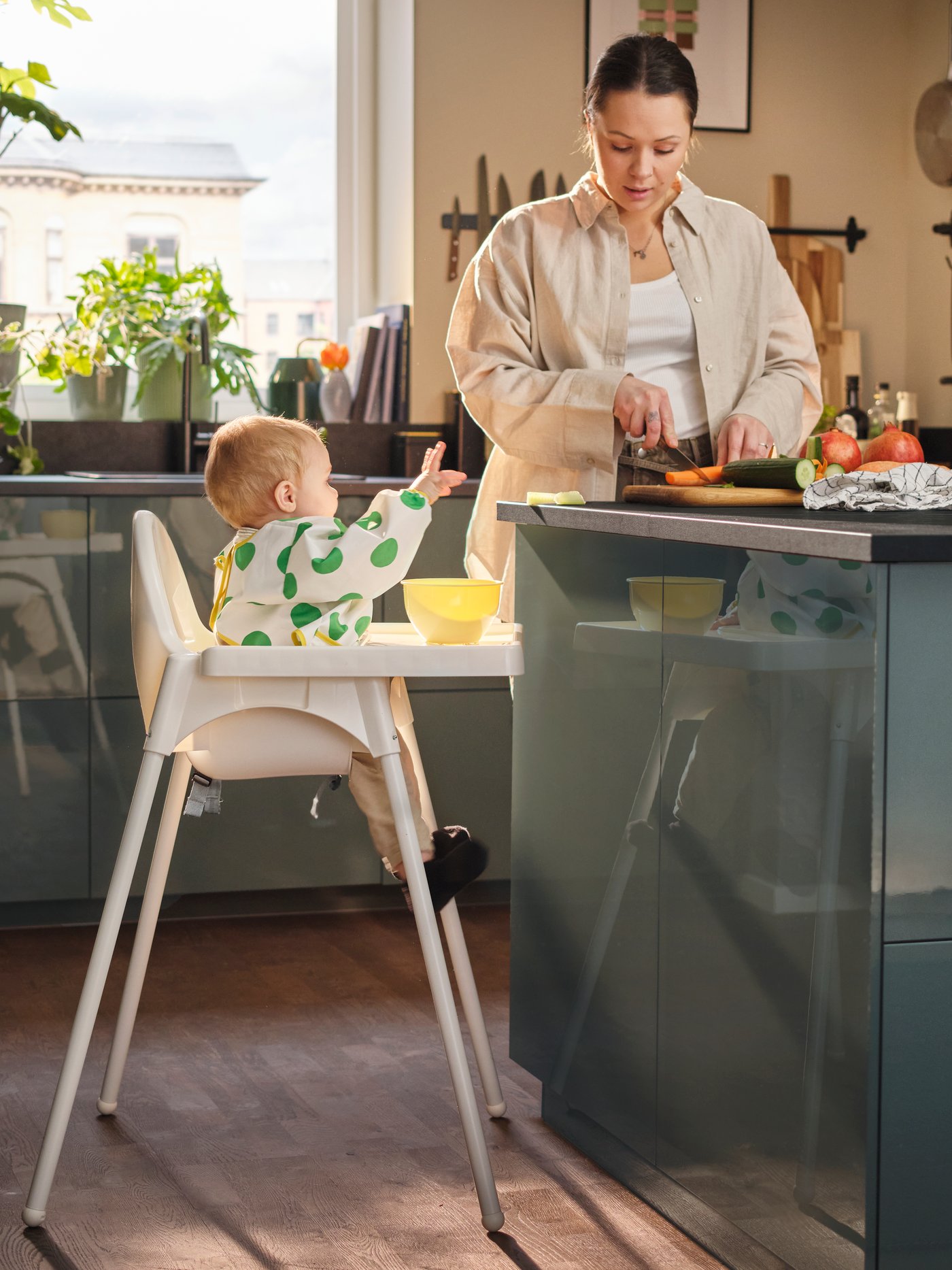 A baby sits in an ANTILOP highchair beside a kitchen worktop wearing a white/green KLADDIG long-sleeved bib and reaching out.