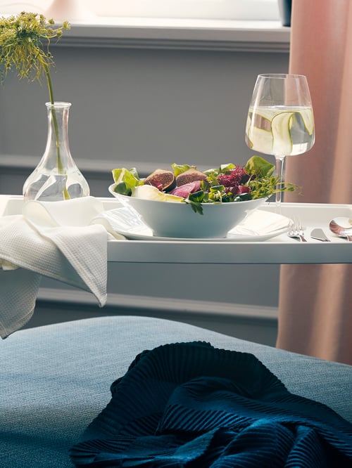 Un plateau de lit KLIPSK blanc est posé sur un canapé. Il contient des couverts, un bol de salade, un verre d’eau et un vase avec une fleur.