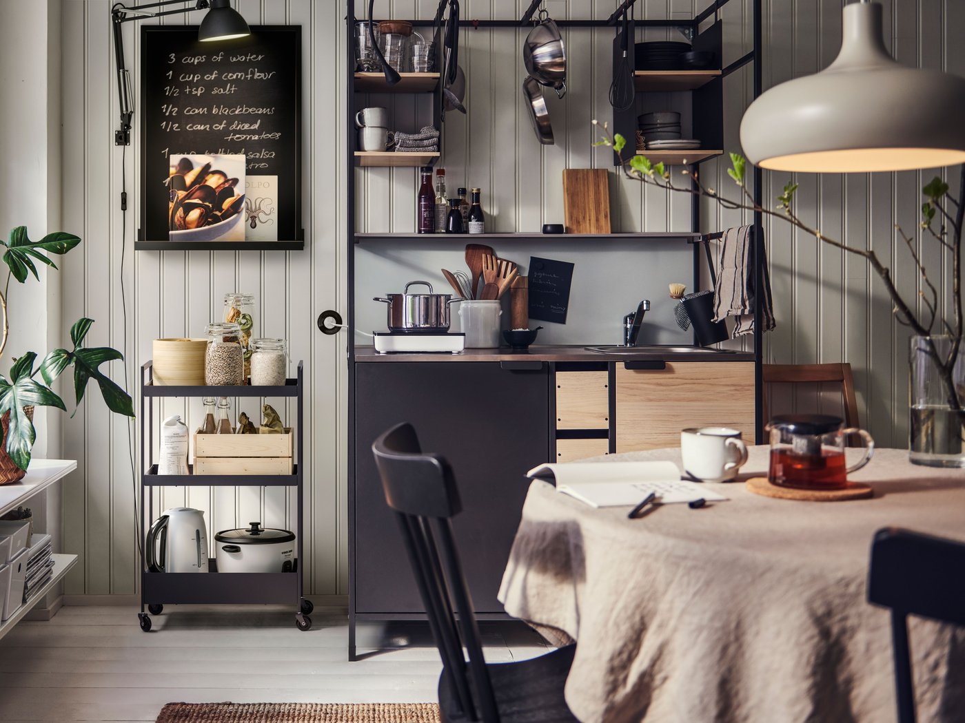 A kitchen with an ÄSPINGE kitchenette, a black NISSAFORS trolley filled with various items, and a dining table with chairs.