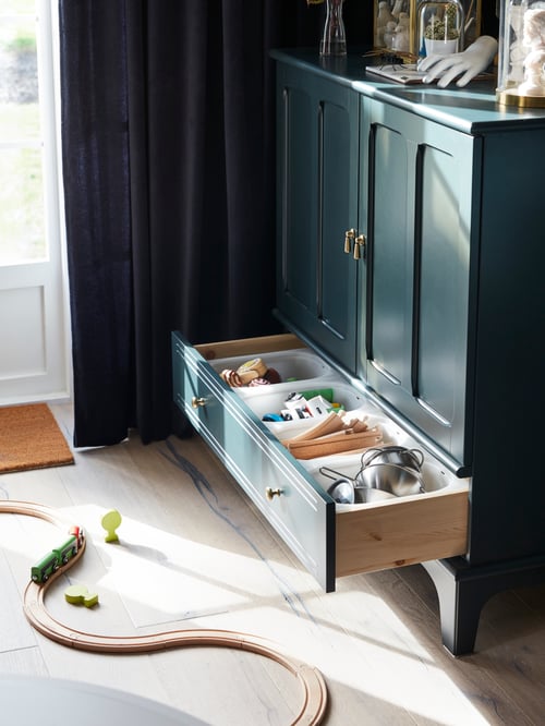 The bottom drawer of a dark blue-green LOMMARP cabinet is open with children’s toys inside. A train set is on the floor.
