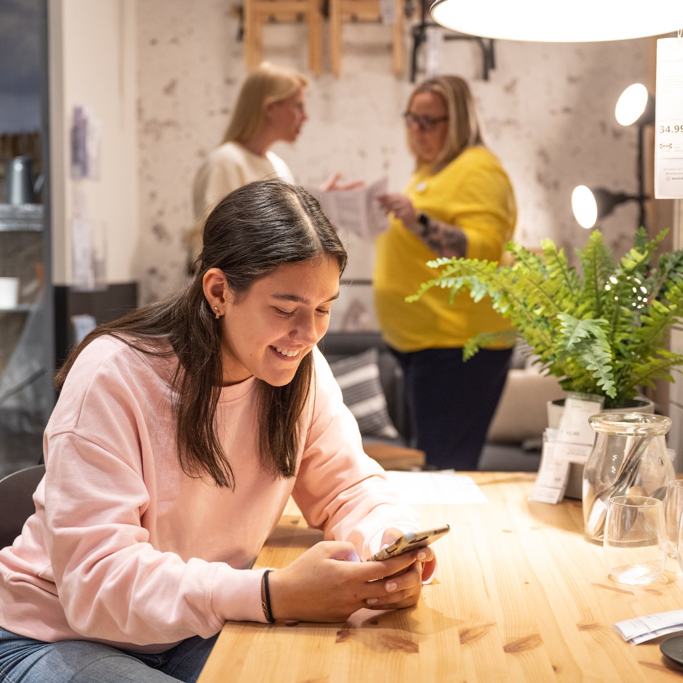 A brown‑haired person in a pink sweatshirt smiles while looking at their phone at a wooden table inside an IKEA store. In the background, an IKEA co‑worker is speaking with a customer.