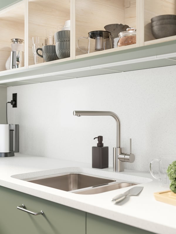 Vista en escorzo de una encimera de cocina de cuarzo blanca sobre un mueble verde oliva, con un lavaplatos empotrado y grifería de cocina cromada monomando. 