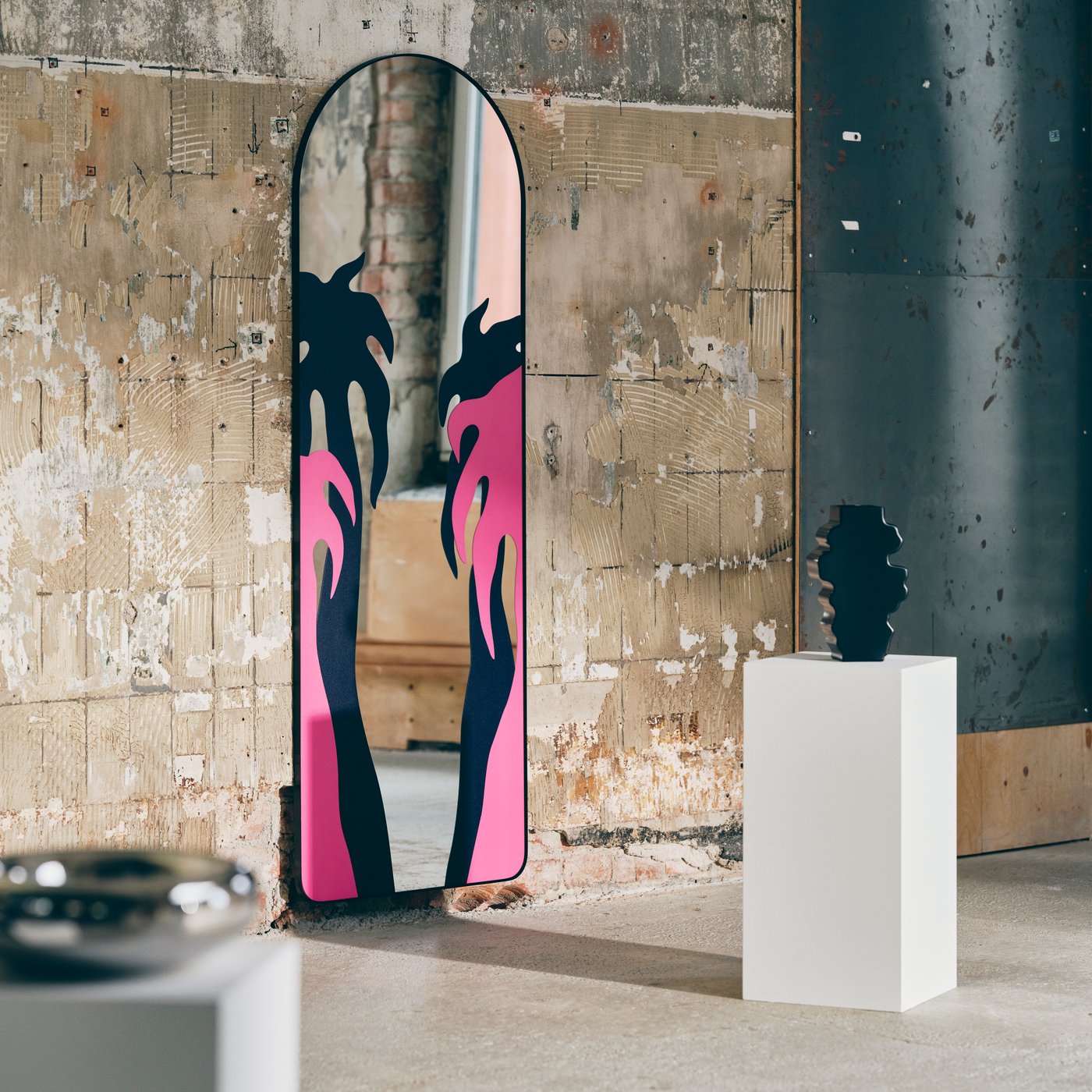 A mirror with pink and black palm tree decoration against a concrete wall, a black sculpture on a white pedestal next to it.