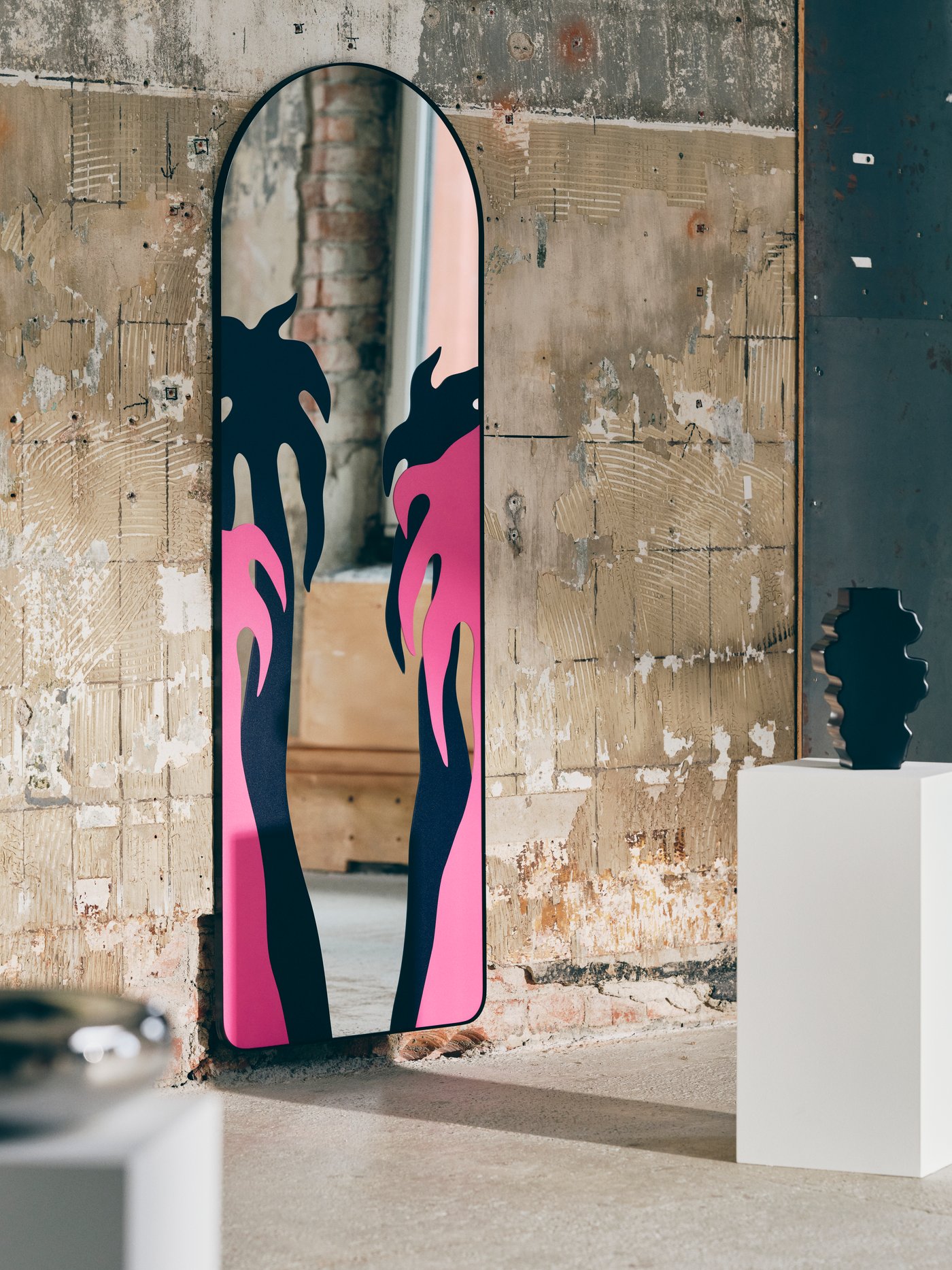 A mirror s pink and black palm tree decoration contra concreta wall, a black sculpture on a white pedestal next to it.