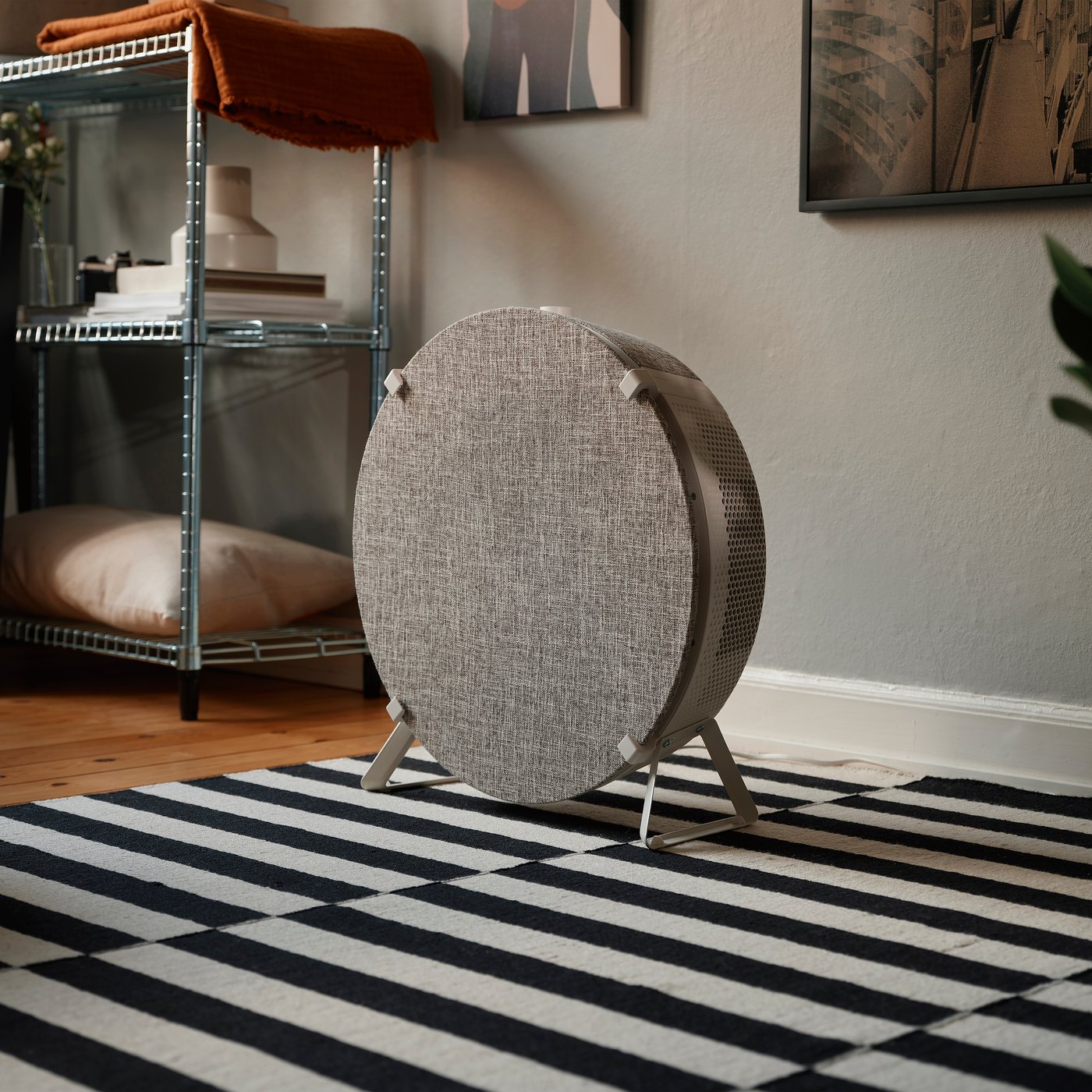 En rund, tygklädd luftrenare står på en randig matta framför en metallhylla med kuddar och en filt.