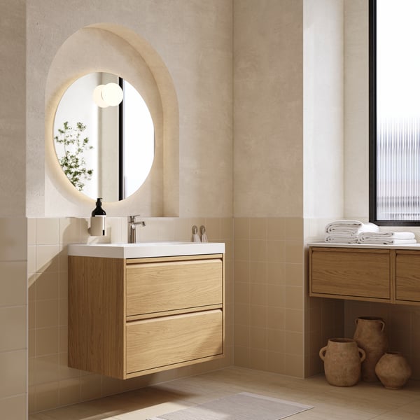 An oak effect, two-drawer ÄNGSJÖN/BACKSJÖN wash-stand is on a wall below a round mirror in a modern beige and white bathroom.
