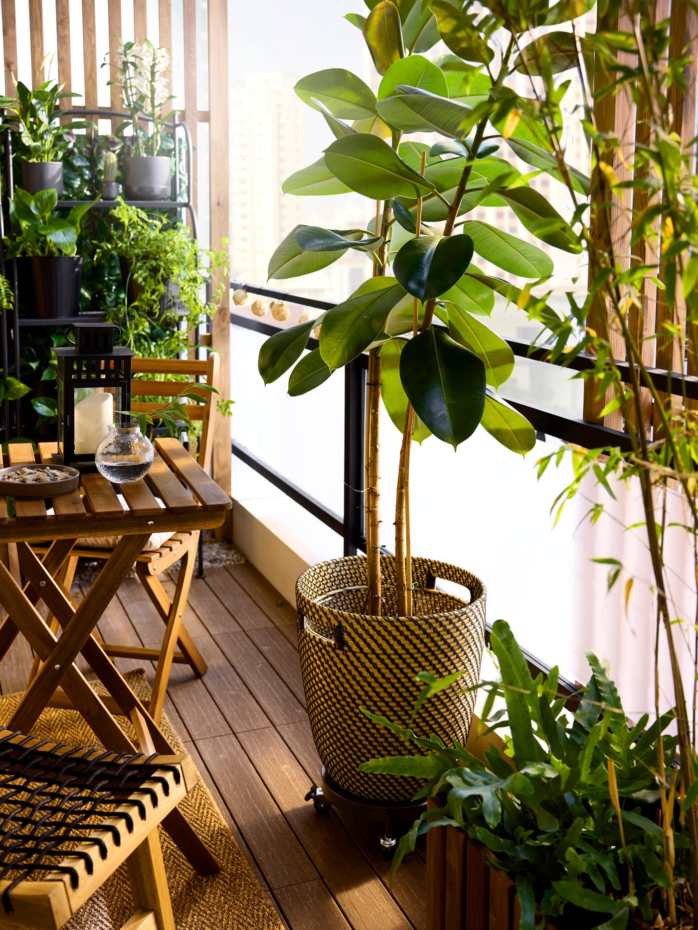 A dark grey/beige woven RÅGKORN in/outdoor plant pot holds a rubber plant on a lush balcony by a glass and metal railing.