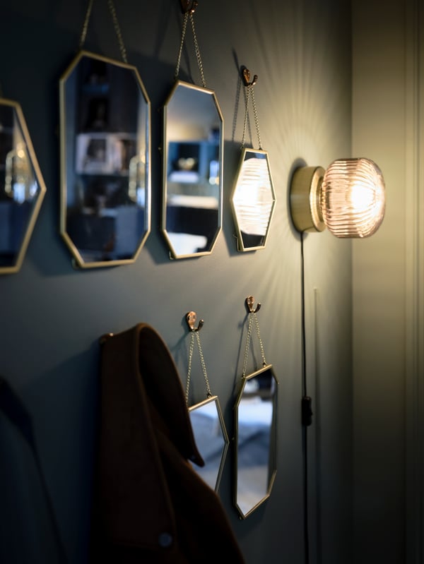 Rows of gold-colour LASSBYN mirrors hang on a green wall amid coats on hooks and a lit wall-mounted lamp.