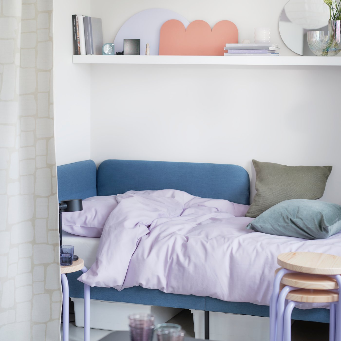 Sovrum med blå soffa som säng, ljuslila sängkläder, och vit vägg-hylla med böcker, dekorationer och växter ovanför.