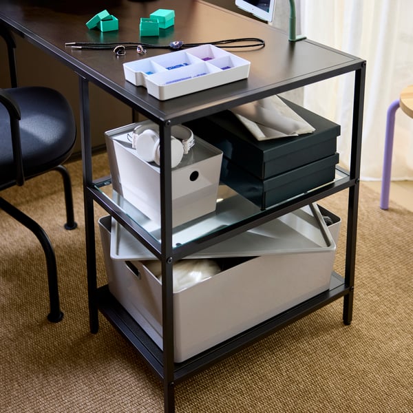 A close-up on a workspace setup with a black-brown VITTSJÖ desk with glass shelves, storing boxes and crafting supplies.