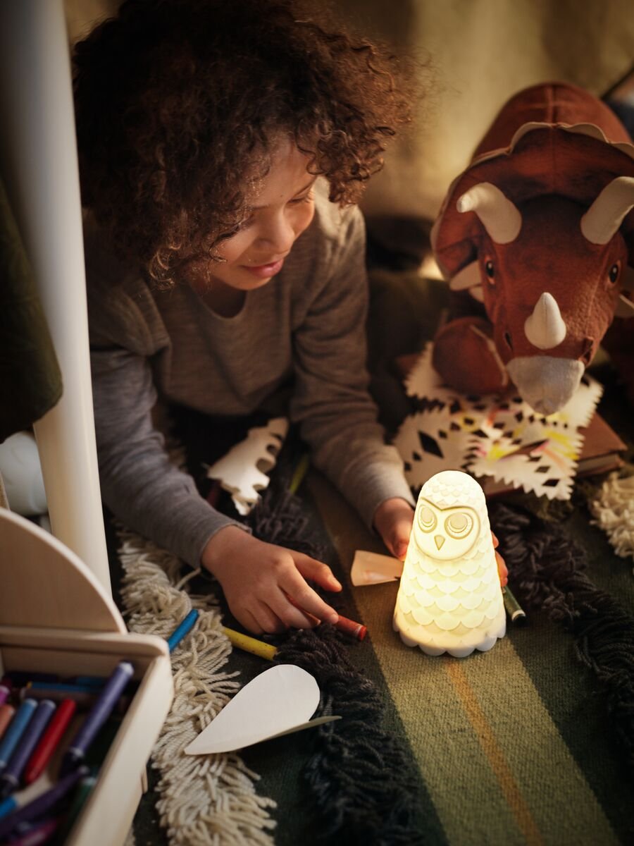 A boy in a cubby made underneath a table looks at a lamp in the shape of an owl. He is surrounded by crayons and paper. A soft toy dinosaur is keeping him company.