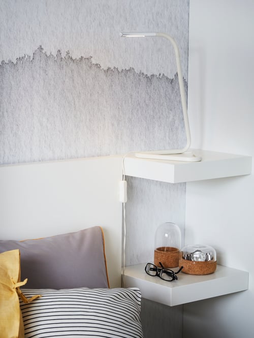 Two, white LACK wall shelves are mounted in the corner of a room by a bed. They hold a lamp, a pair of glasses and ornaments.