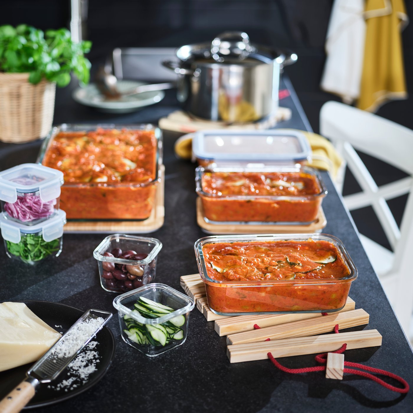 A kitchen setting features glass containers of lasagna, arranged with fresh ingredients and utensils.