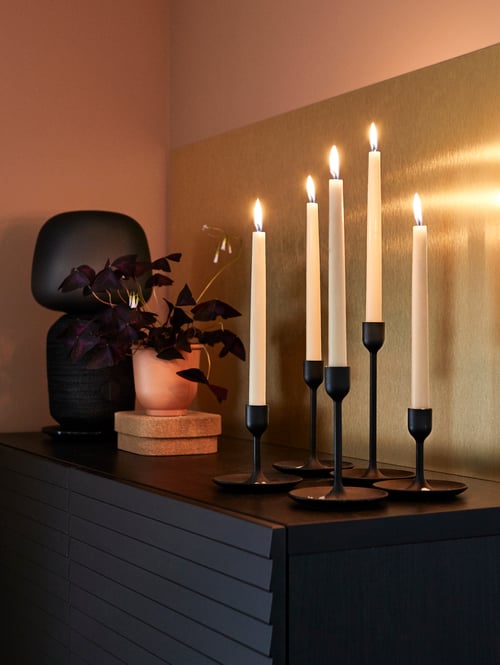 Living room with black chest of drawers, brass wall panel, table lamp, and five candlesticks with white lit candles.