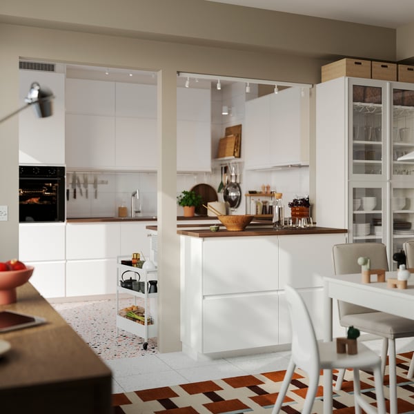 A bright kitchen has matt white VOXTORP fronts and a kitchen peninsula with a white dining table in the foreground.