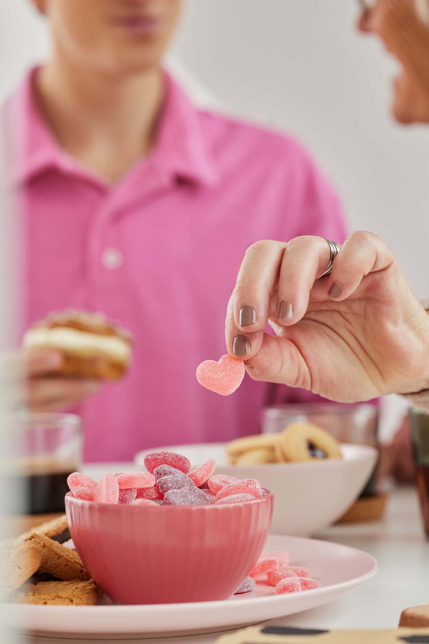En hånd tager en hjerteformet vingummi fra en skål, mes der er to personer i baggrunden. 