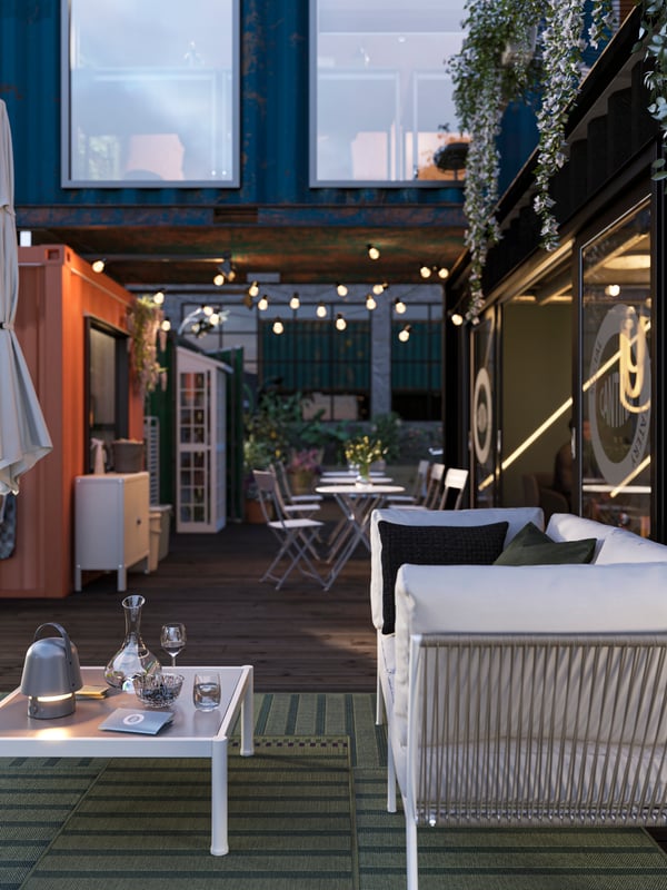 An outdoor seating area with a SEGERÖN sofa and bistro sets, is lit up by a VAPPEBY LED speaker lamp and lighting chains.