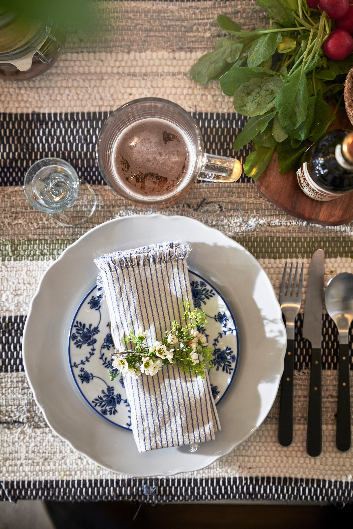 
Ovanifrån‑vy av en dukning med vit tallrik på en randig textilservett, dekorerad med ett snöre och små blommor; bredvid ligger bestick och ovanför syns en kopp samt grön växtdekoration på ett rustikt bord.
