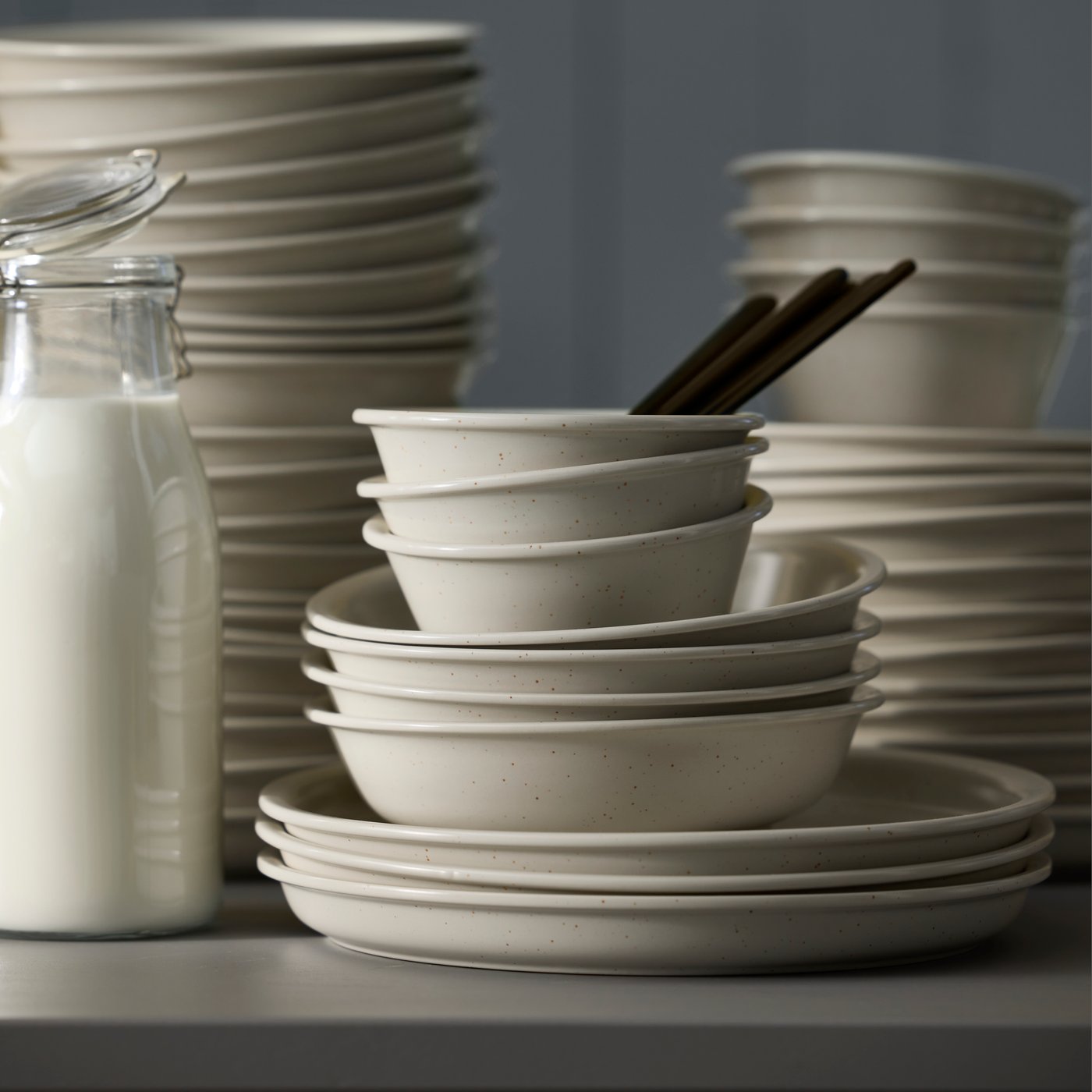 Several light beige DAGGASTER bowls, plates and deep plates are stacked on a table next to a glass jar of milk.