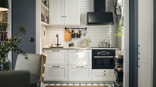 A white-tiled kitchen with matte white AXSTAD fronts and tall cabinets has black appliances and shelves on the wall.