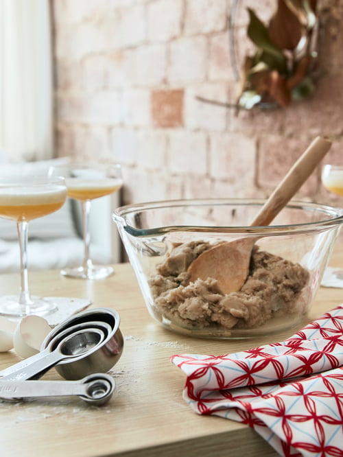 Kitchen with brick wall, glass bowl with dough, wooden mixing spoon, set of five measuring cups, champagne coupes, tea towel.
