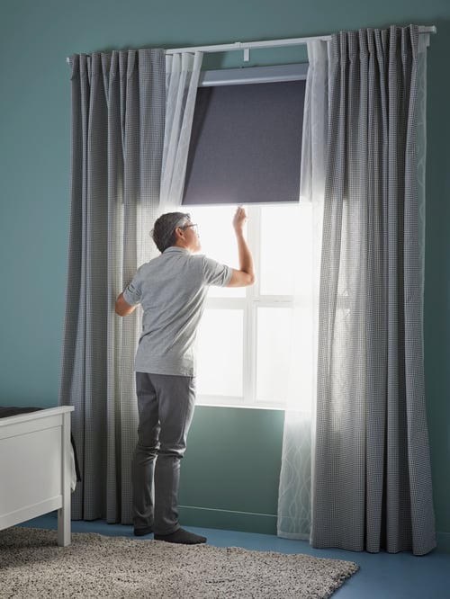 A man is standing by a bedroom window with layered curtains. He is pulling down a light grey TRETUR block-out roller blind.