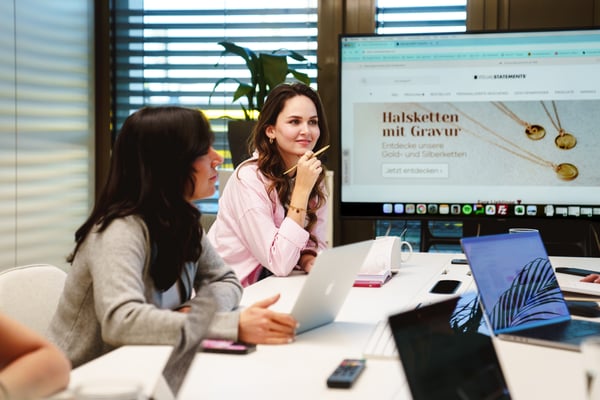 Personen sitzen an einem Konferenztisch mit Laptops und einer Präsentation auf einem Bildschirm im Hintergrund.
