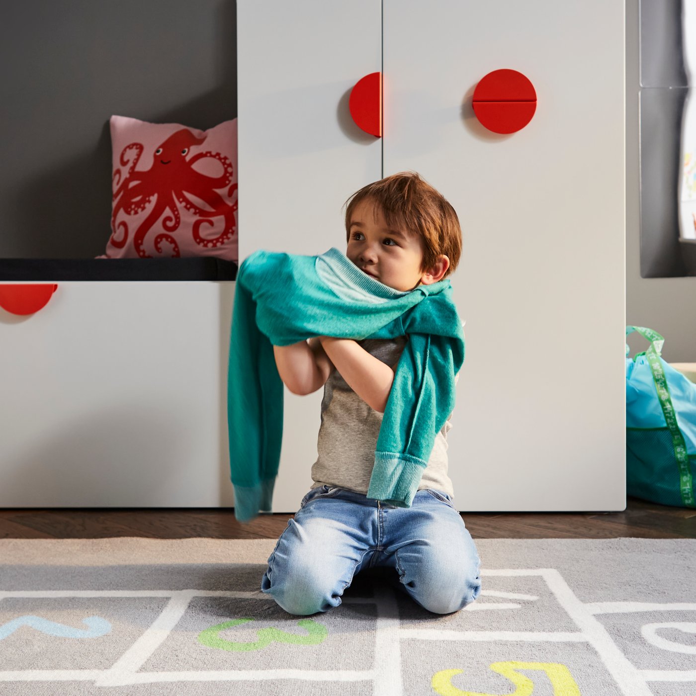 A child getting dressed on a grey rug in front of a white bench with toy storage and a white wardrobe with a pull-out unit.