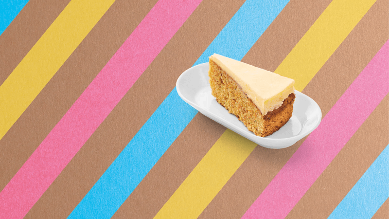 Ein Stück Möhrentorte auf einem weißen Teller, der auf einem bunt gestreiften Untergrund steht.