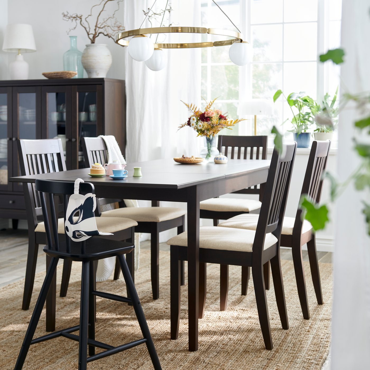 A dark brown NÄSINGE extendable table is set with chairs in a bright dining area with large windows and decorative plants.