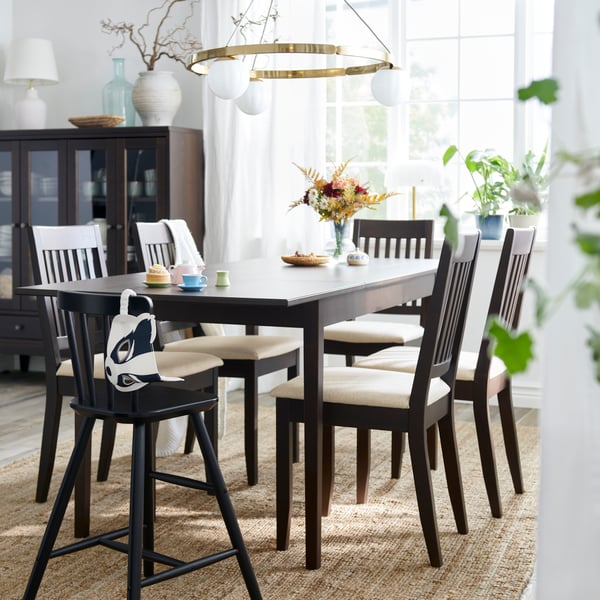 A dark brown NÄSINGE extendable table is set with chairs in a bright dining area with large windows and decorative plants.