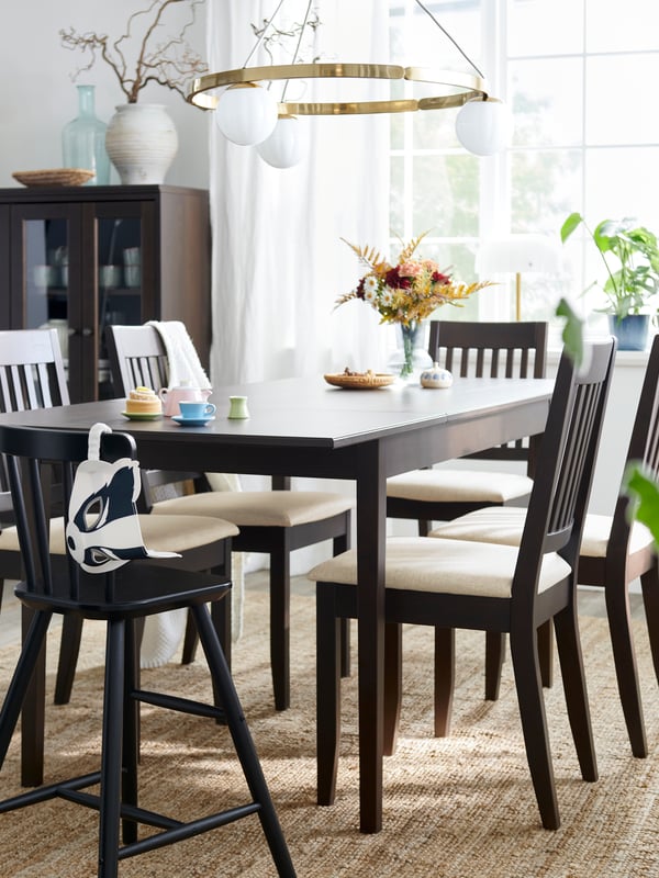 A dark brown NÄSINGE extendable table is set with chairs in a bright dining area with large windows and decorative plants. 