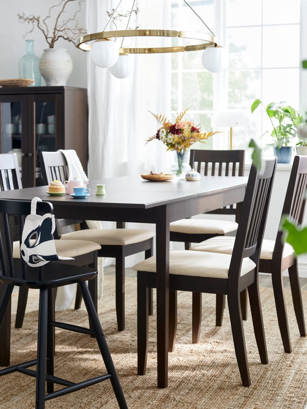 A dark brown NÄSINGE extendable table is set with chairs in a bright dining area with large windows and decorative plants. 