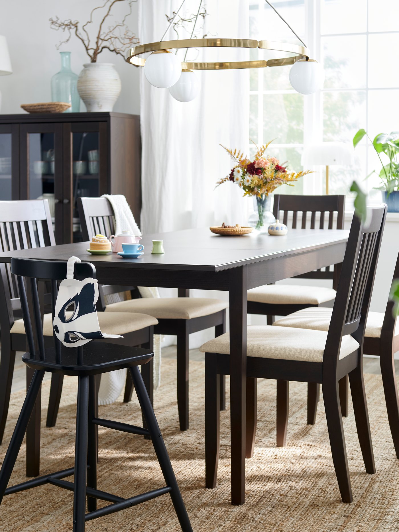 Ett mörkbrunt NÄSINGE förlängningsbart bord är placerat med stolar i en ljus matplats med stora fönster och dekorativa växter.