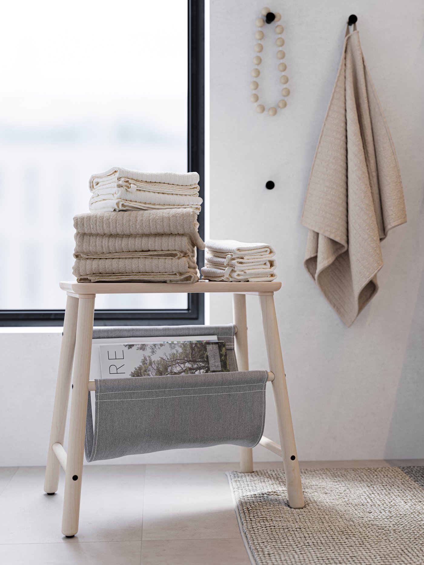 A wooden stool with neatly folded beige and white towels on top. Black hooks with a beige towel and wooden necklace.