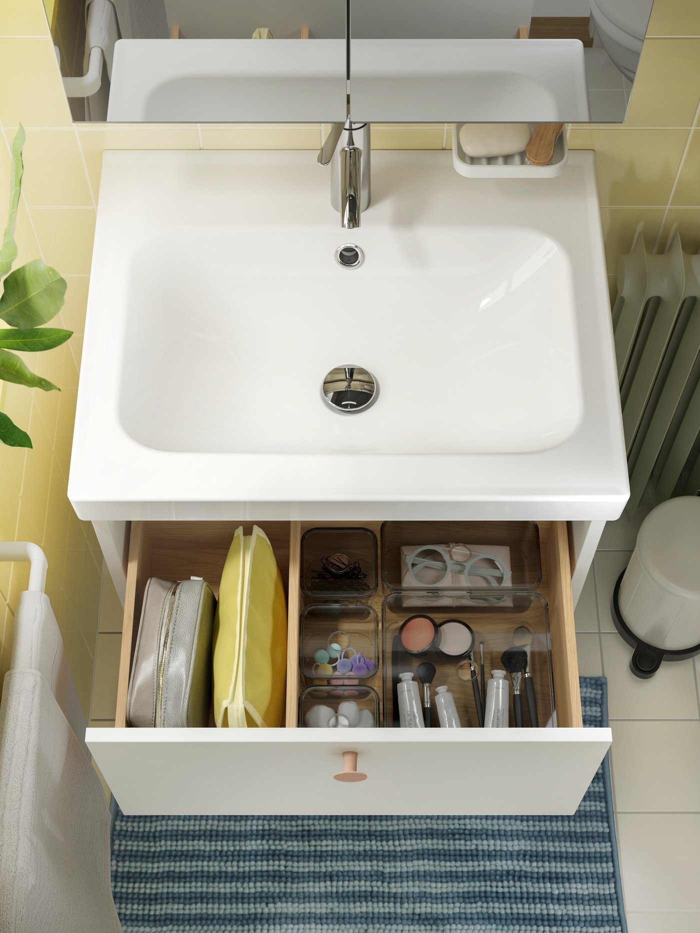 Een badkamermeubel met open lade toont een overzichtelijke indeling van toiletartikelen, make-up en verzorgingsproducten, met een spiegel erboven en een blauw gestreept tapijt ervoor.