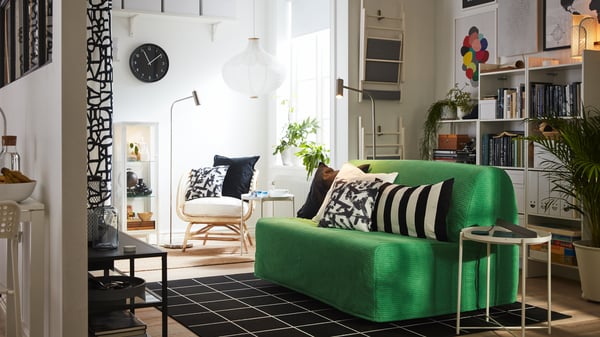 Ein LYCKSELE LÖVÅS 2er-Bettsofa mit einem leuchtend grünen Bezug steht in der Mitte eines kleinen Raums. Daneben ein Beistelltisch und dahinter weiße Regale. 