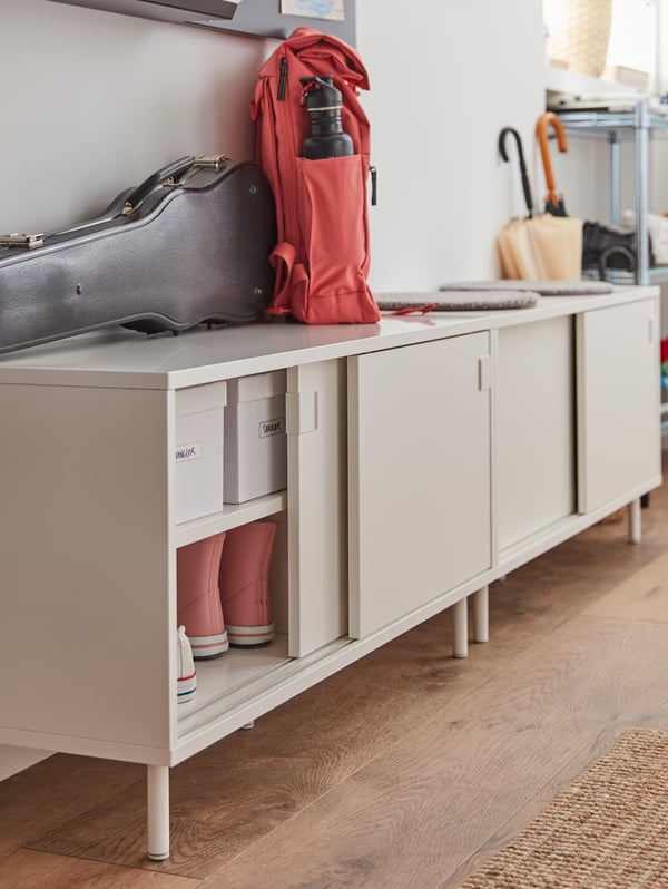 A white bench with sliding doors reveal white boxes and sneakers in them. A violin case and a red backpack is on top.