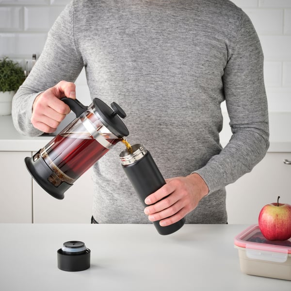 A person’s torso is shown wearing a grey sweater, holding a French press coffee maker with freshly brewed coffee. The French press is being tilted to pour coffee into a cup, which is not visible in the frame. The kitchen background features modern, clean lines with a white countertop and cabinets.