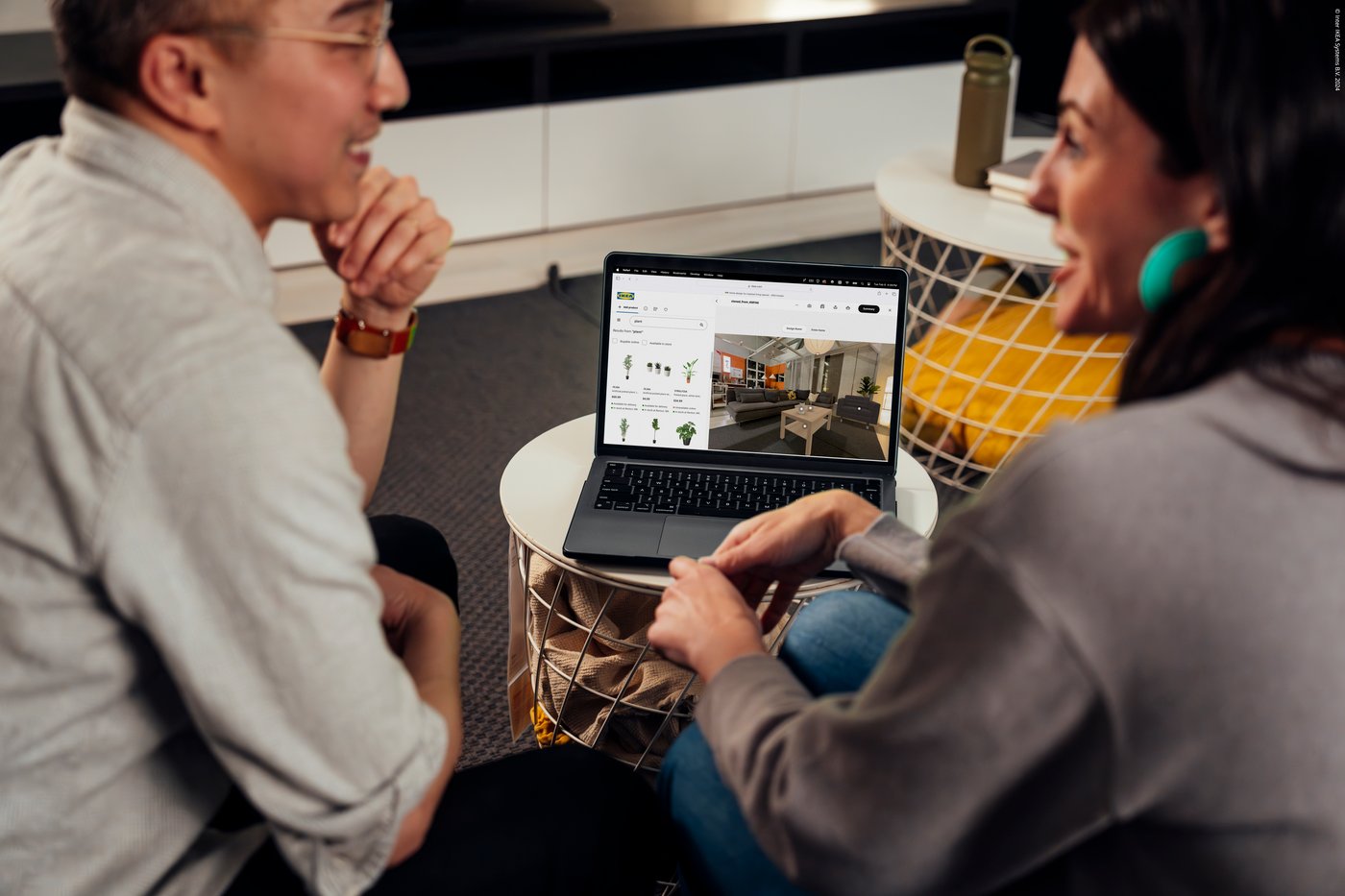 Två personer sitter vid ett bord och tittar på en laptop som visar en sida med inredning, i en hemmiljö med soffa och kuddar i bakgrunden.
