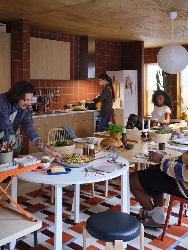 Vier mensen bereiden een maaltijd voor in een levendige keuken. Eén persoon leunt over een witte VIHALS tafel en legt er eten op.