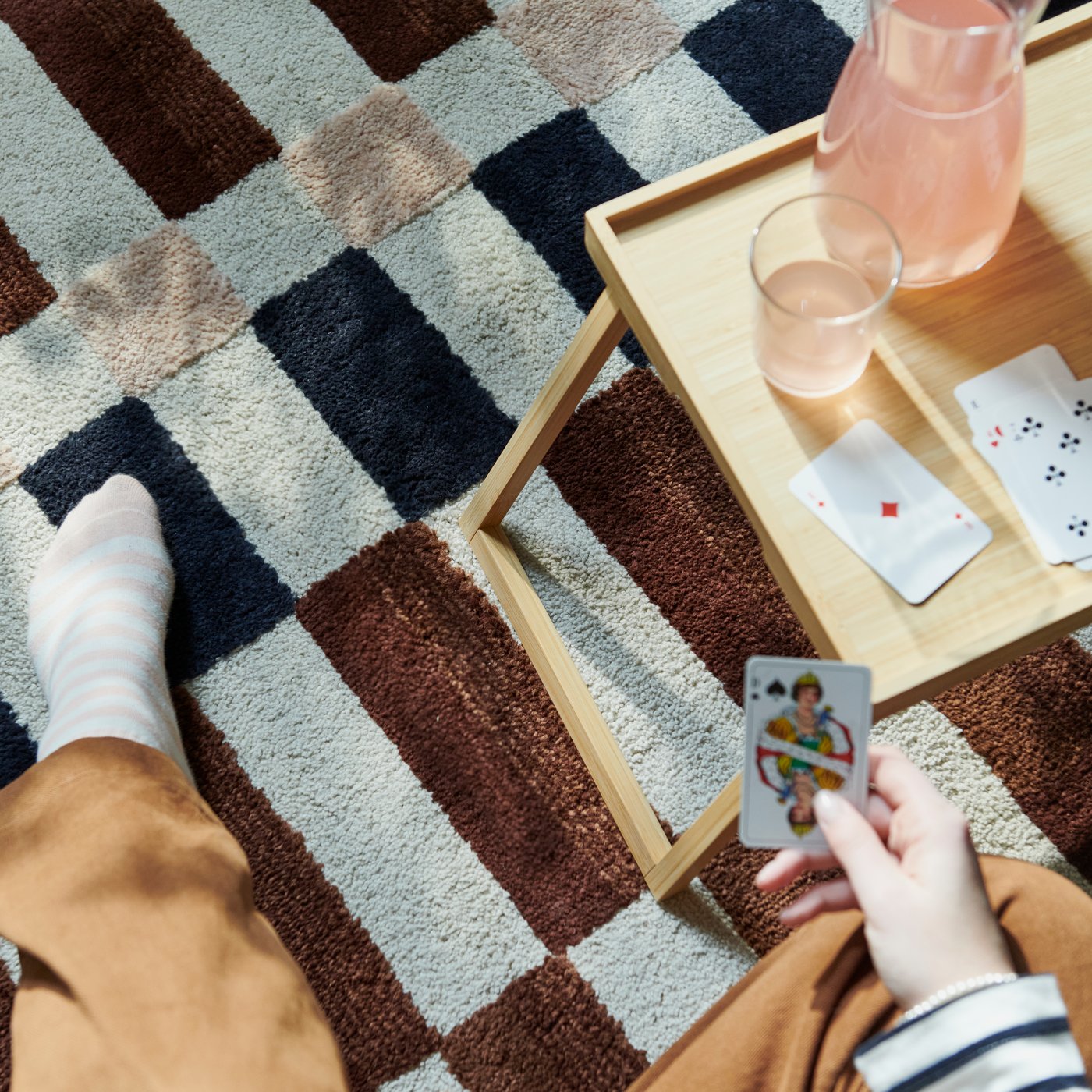A small wooden table with folding legs sits on top a checkered MARKBLOMME rug, while a person is playing cards.