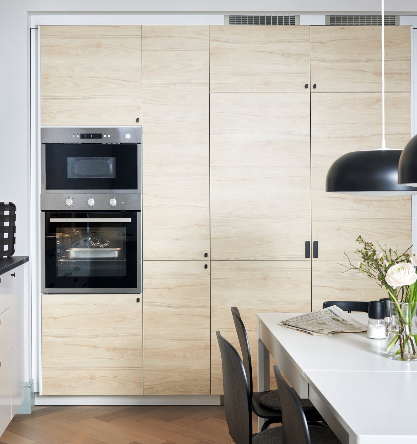 Et lyst køkken, hvor man ser højskabe af træ og aner et spisebord i forgrunden.