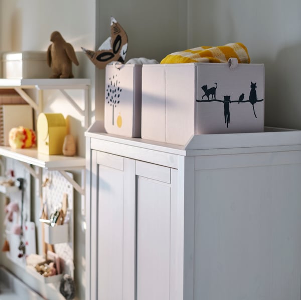 Unha caixa BARNDRÖM beixe á vista na parte superior dun armario SUNDVIK branco con obxectos decorativos ao redor nun espazo acolledor.