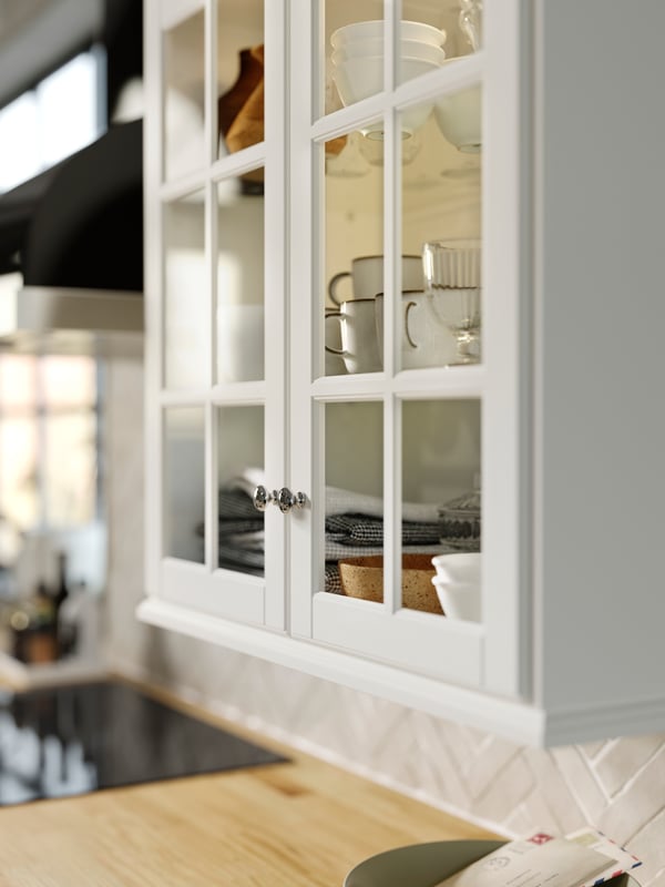 Une armoire BODBYN blanc cassé avec des portes en verre et des tablettes sur lesquelles se trouvent des tasses GLADELIG grises et des bols VARDAGEN blanc cassé.