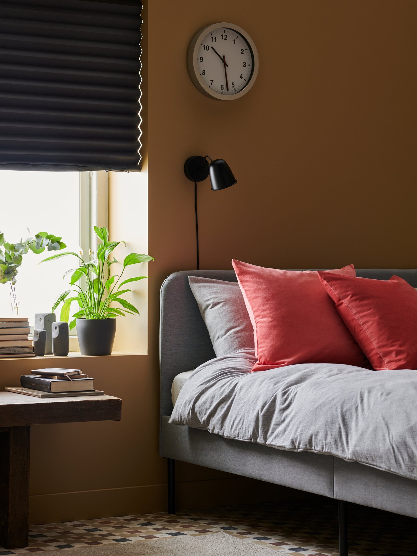 Cama junto a una ventana con un estor SCHOTTIS de color gris oscuro. En el alféizar hay libros y una planta en un macetero PERSILLADE.