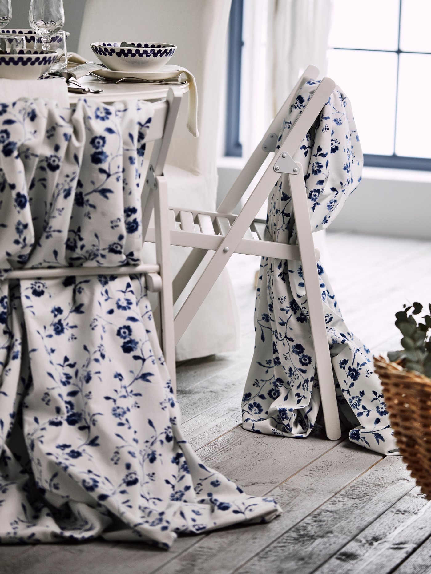 Two white folding chairs draped with white and blue flower patterned BLÅGRAN fabric, standing next to a festive table.