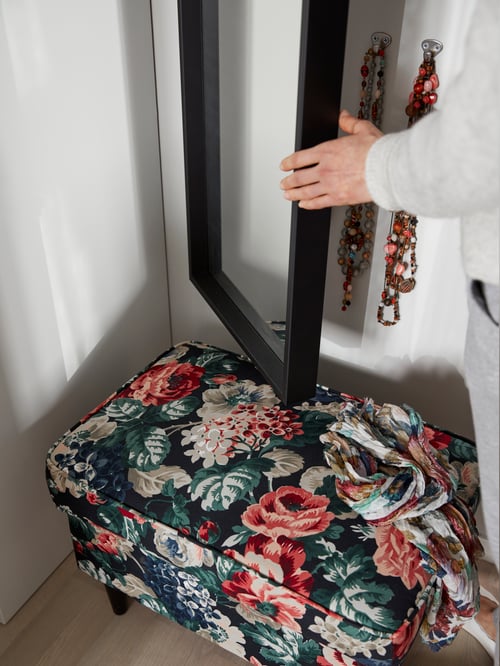 O poză de aproape a unui bărbat atingând rama neagră a unei oglinzi care stă în fața unui taburet cu model floral.