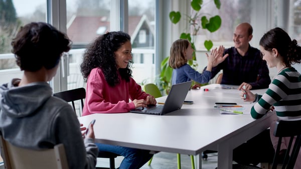 A group of four people sitting around a table in a bright room with large windows. One woman in a pink sweater is working on a laptop and smiling, while another person in a striped sweater is engaged in a conversation