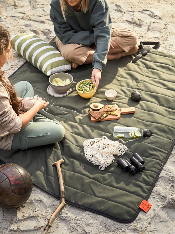 Dos mujeres sentadas sobre una manta en la playa haciendo un picnic. Sobre la manta se ven envases para alimentos con frutas. También hay una tabla para picar sobre la manta con un pan de molde integral y al costado una botella de vidrio con jugo.